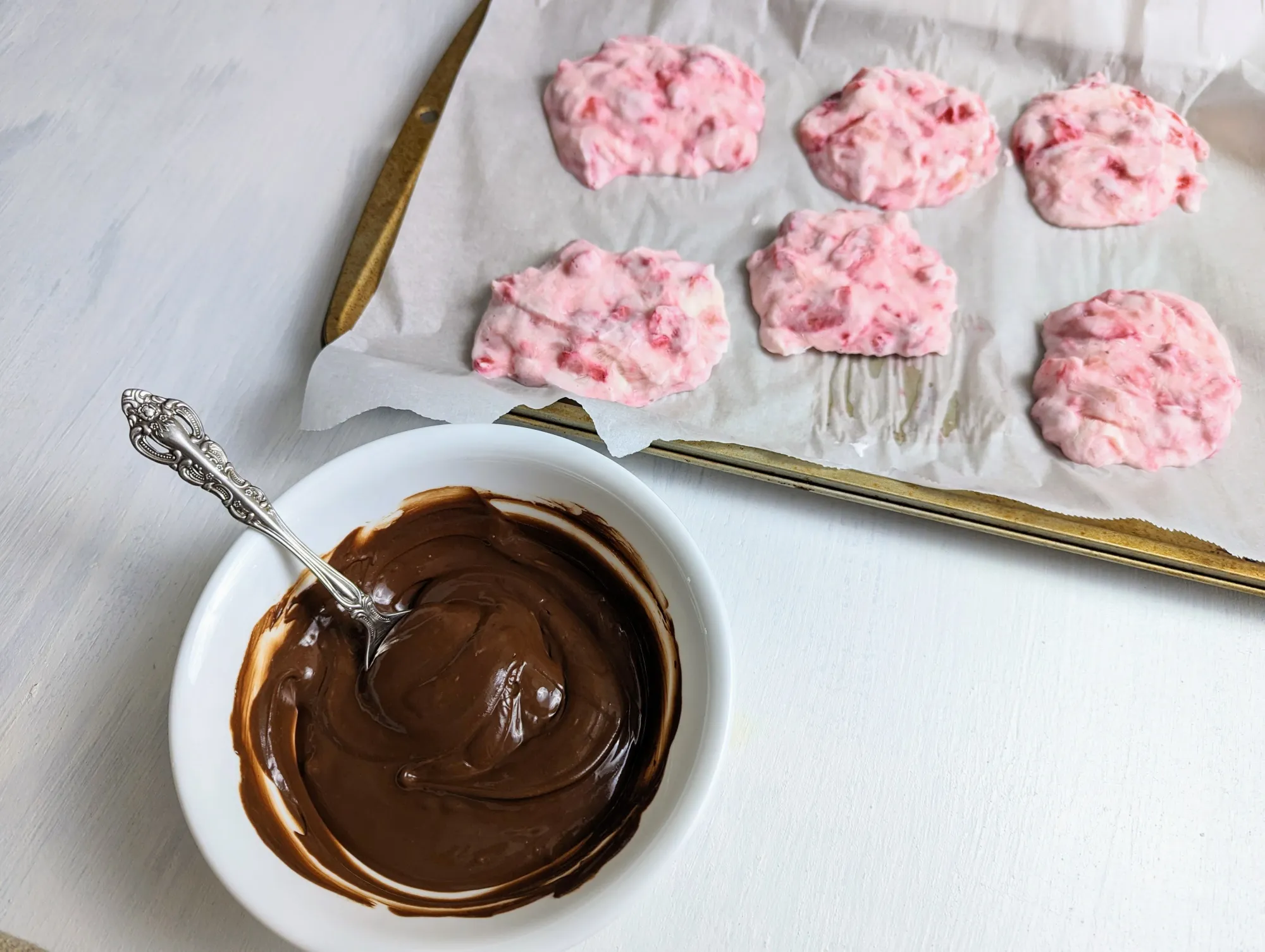 A bowl of melted chocolate with a spoon, next to a tray of frozen lumps of greek yogurt and strawberries