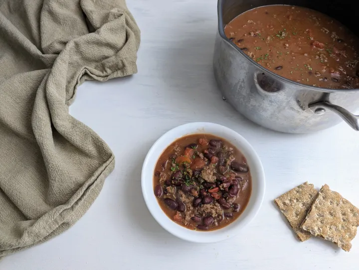 Chili in a white bowl, with some crackers and a pot of chili next to it