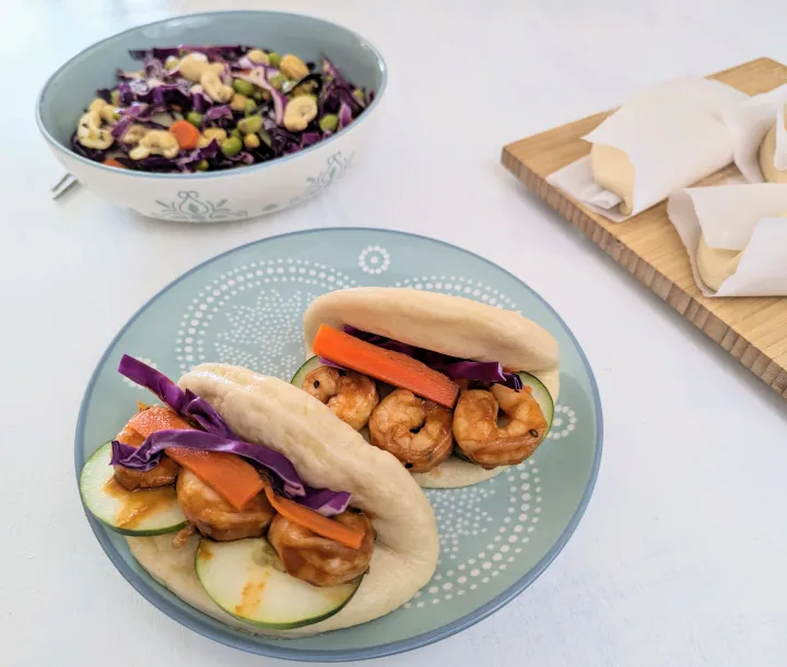 A shrimp bao bun on a plate, filled with cabbage, cucumber and carrot