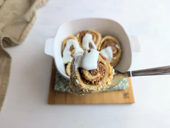 A cinnamon roll on a serving spoon above a dish of more cinnamon rolls with white frosting